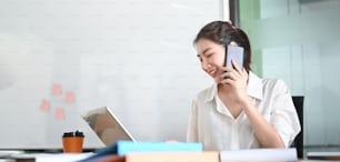A businesswoman is talking on the mobile phone while sitting and using a computer laptop at the working desk.