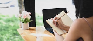 A beautiful woman is taking notes while sitting in front of a computer laptop at the wooden working table.
