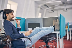 Fröhlicher männlicher Arbeiter, der ein Glas alkoholisches Getränk hält und lächelt, während er im Büro eine Zigarre genießt