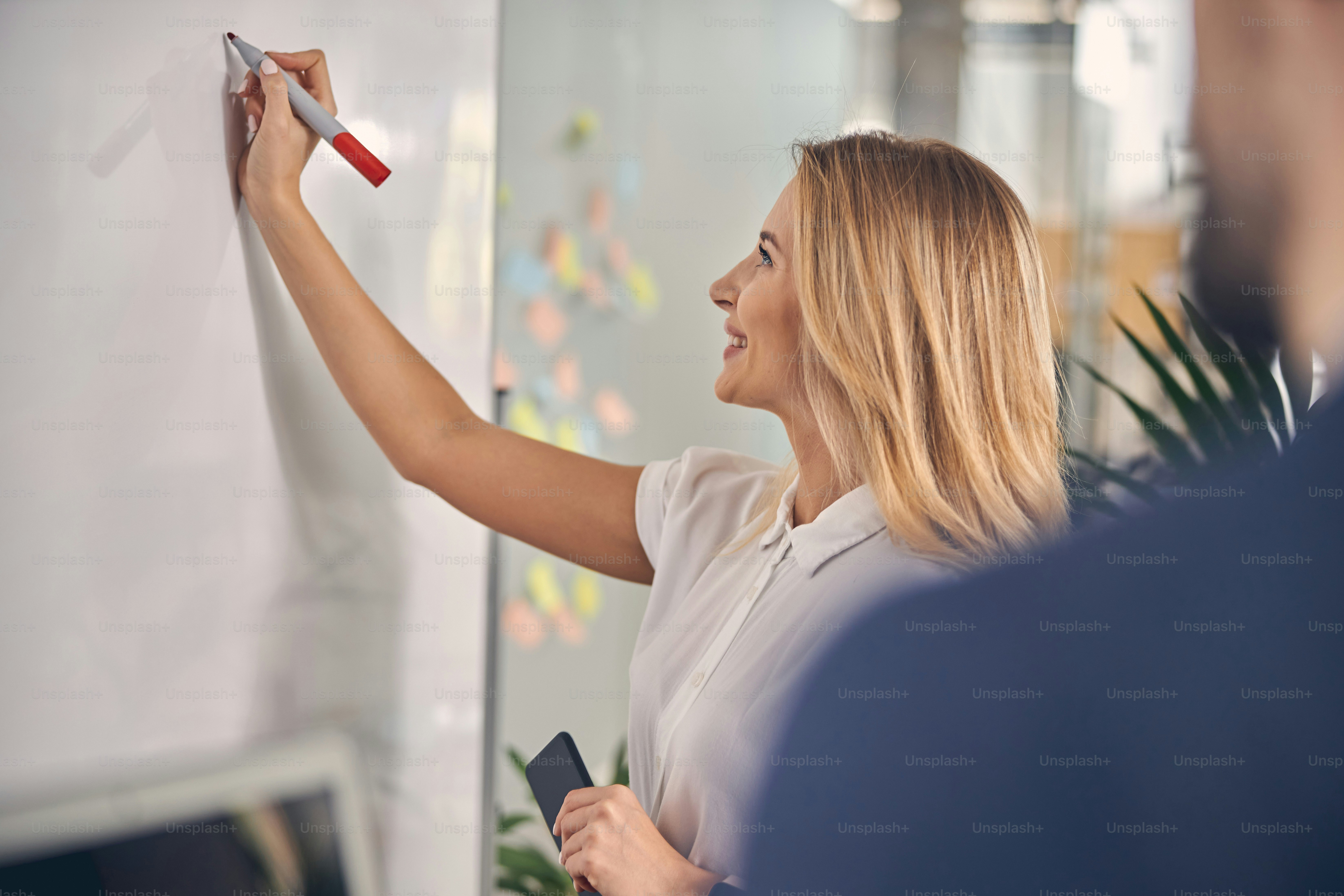 Schöne blonde Dame, die Notizen auf Bürobrett macht und lächelt, während sie das Smartphone hält