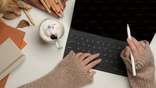 Overhead shot of female using digital tablet with keyboard on worktable with stationery and cup of beverage