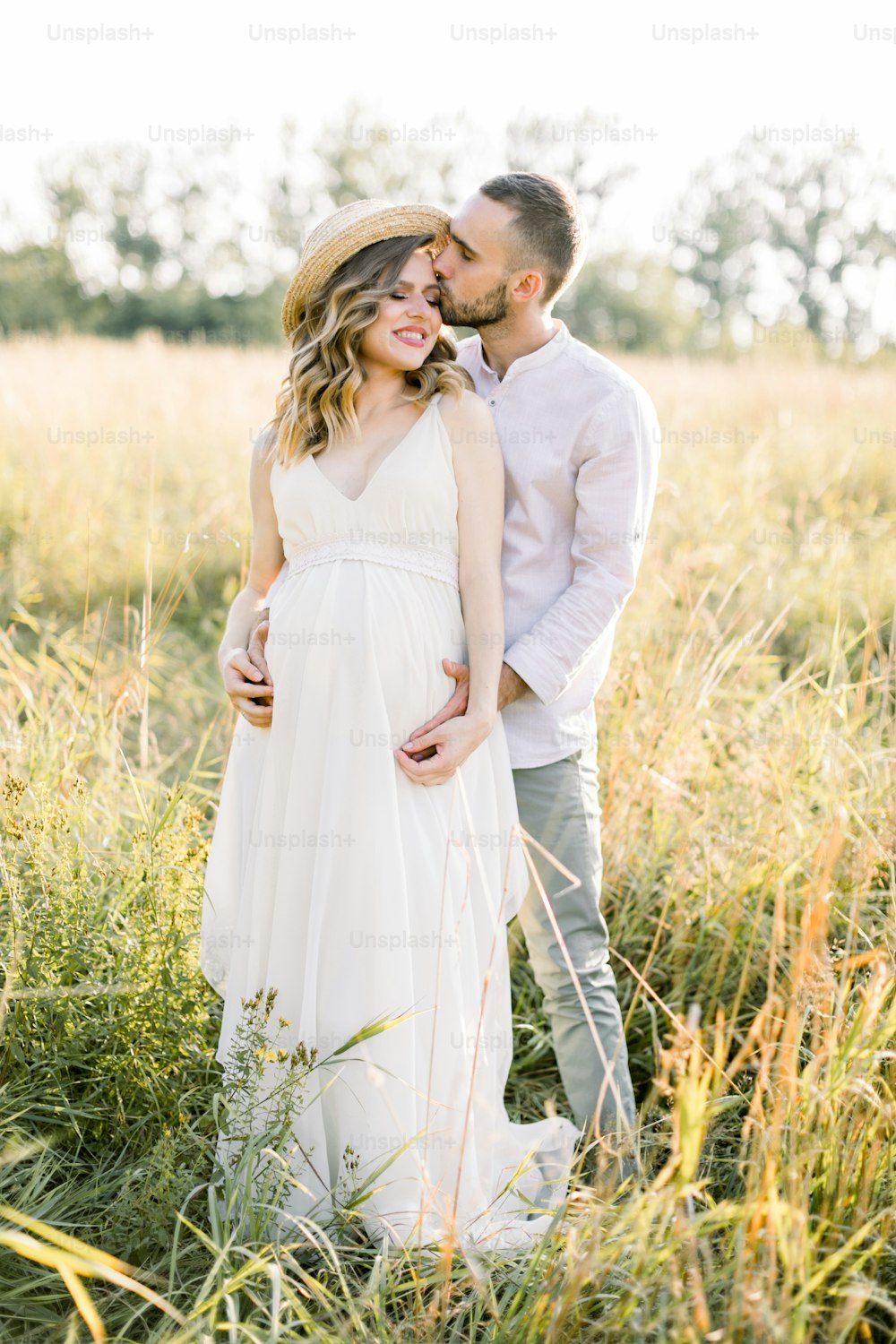 Young beautiful pregnant couple in white clothes walking in a field in the summer at sunset. Handsome bearded young man kissing his pretty pregnant lady in white dress and straw hat.