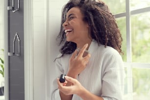 Cheerful pretty young female is laughing while spraying perfume on body from bottle at home