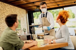 Happy waiter serving coffee to a couple while wearing face mask during coronavirus epidemic.