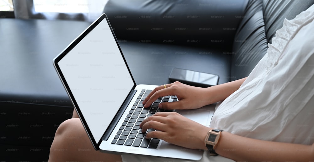 La imagen recortada de una mujer está usando una computadora portátil con pantalla blanca en blanco mientras está sentada en el sofá.