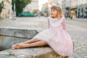 Ritratto romantico di affascinante bambina caucasica in abito da principessa rosa alla moda e senza scarpe, seduta sulla vecchia fontana di pietra sulla strada sullo sfondo della città antica.