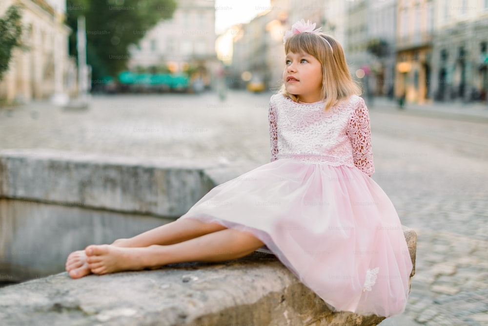 Romantic portrait of charming Caucasian little girl in trendy pink princess dress and without shoes, sitting on the old stone fountain on the street on ancient city background.