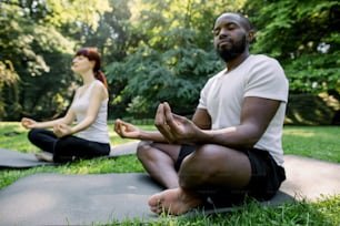 Close up of young African man sitting in lotus yoga position, zen gesturing, outdoors in nature. Pretty woman meditating behind. Concept of healthy physically and mentally lifestyle and relaxation