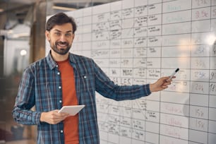 Handsome young man looking at camera and smiling while standing by planner board and holding electronic pad PC