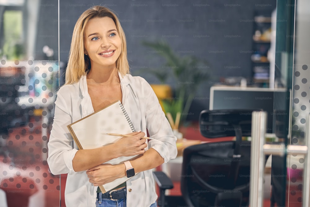 Beautiful female worker looking away and smiling while holding spiral sketchbook