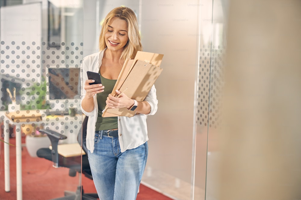 Charming blonde lady checking messages on smartphone and smiling while holding documents