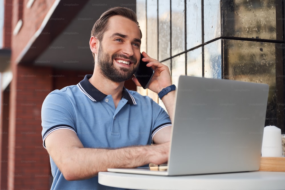 Low angle of smiling bearded male standing at table and using laptop while talking on smartphone