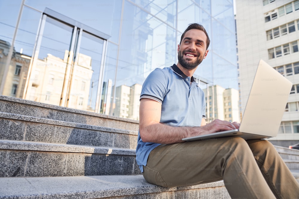 Low angle of smiling young male sitting on steps in city centre and working with notebook