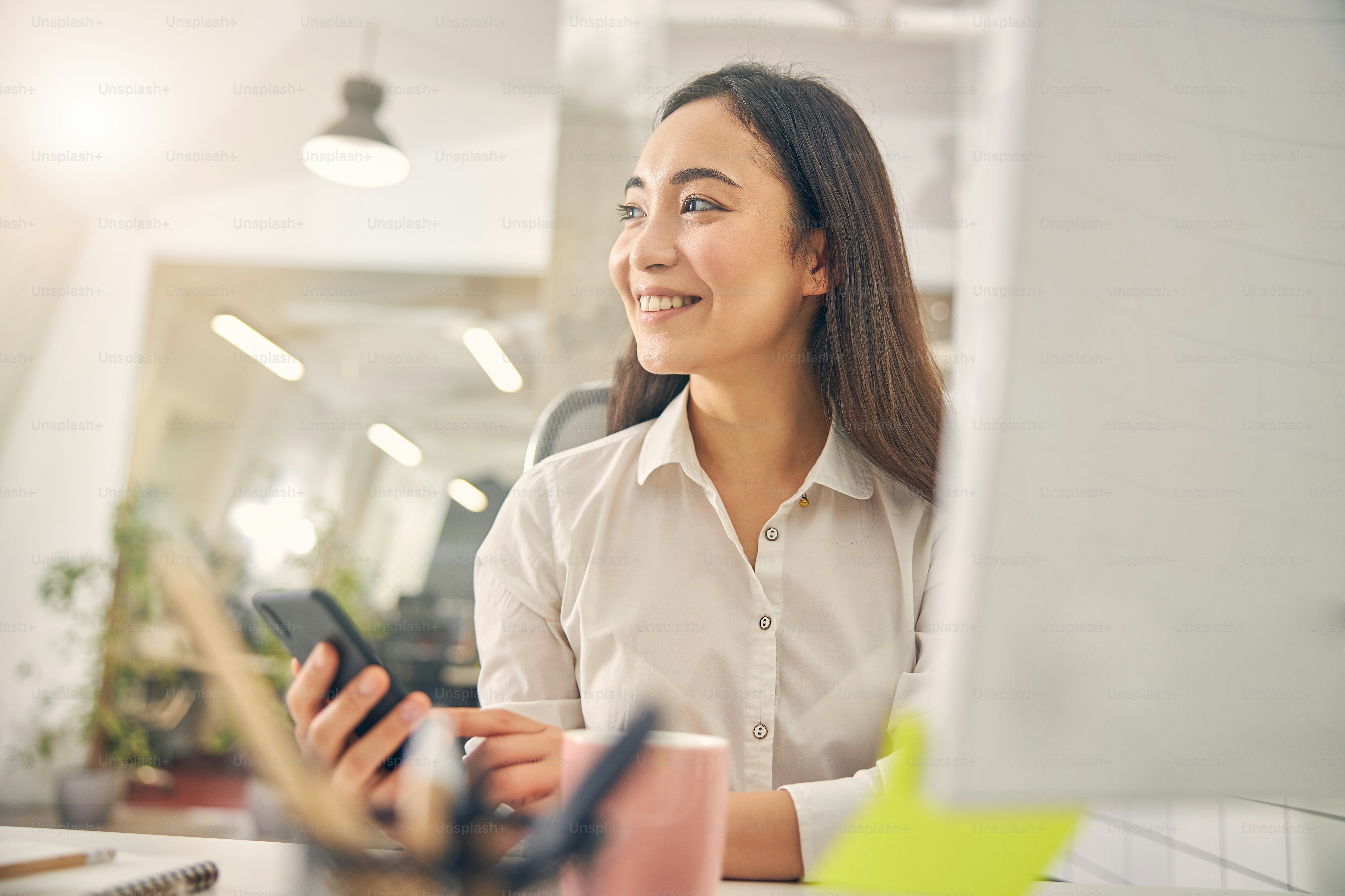 Jolie femme brune gardant le sourire sur son visage tout en étant assise sur son lieu de travail, regardant de côté