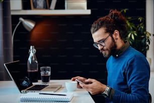 Side view of serious hipster sitting in his office and using smart phone.