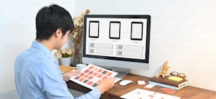 Un diseñador gráfico está seleccionando un color mientras está sentado frente a una computadora en la mesa de madera.