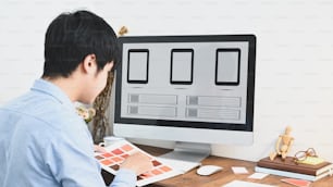 A graphic designer is selecting a color while sitting in front of a computer at the wooden table.