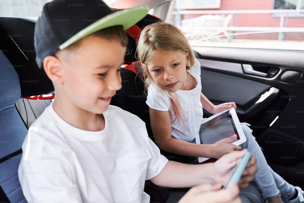 Menino alegre está usando smartphone enquanto sua irmã está se divertindo com tablet durante a viagem