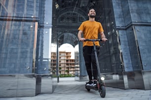 Low angle of happy handsome bearded male riding powered ecological transport in glass urban arch