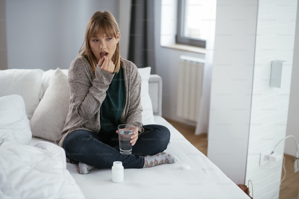 Young sick woman taking medicine pill. Beautiful woman sitting on sofa having stomach pain.