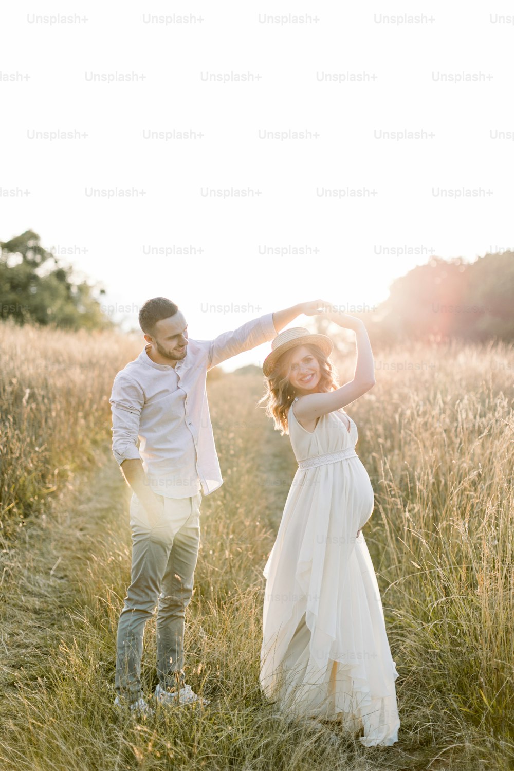 Jeune couple caucasien amoureux marchant et dansant dans un beau champ d’été. Femme enceinte en chapeau et robe blanche dansant avec son bel homme. et concept de grossesse