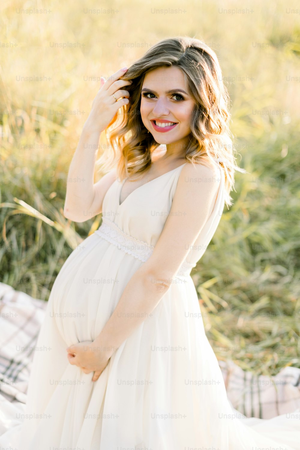 Pretty caucasian pregnant woman in white dress sitting on a picnic blanket on a meadow at sunset. Summer evening in nature. Happy maternity and pregnancy concept.