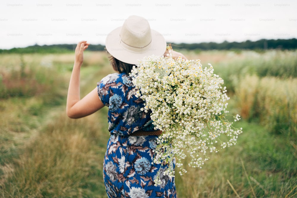 田舎の夏。青いビンテージドレスと帽子をかぶったスタイリッシュな若い女性が、夏の牧草地で白い野の花と一緒に歩いています。フィールドで大きなヒナギクの花束を持つ美しい女の子、後ろ姿。
