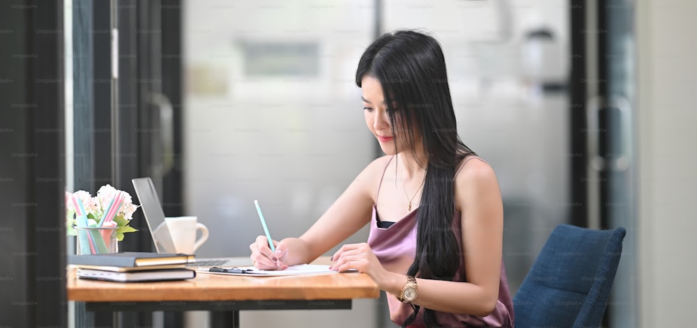A beautiful woman is taking notes at the wooden working table.