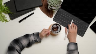 作業台にコーヒーカップを持ちながら、キーボードでデジタルタブレットで作業する女性サラリー�マンの俯瞰ショット