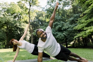 Multiethnic joyful young man and woman exercising in a park, holding side-plank position with one arm raised up. Fit couple training outdoors in the early morning sunshine