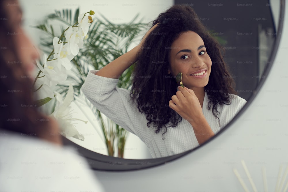 Mulher jovem cacheada feliz está fazendo massagem facial com gadget cosmetologia antes do espelho no banheiro em casa