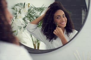 La giovane donna riccia felice sta facendo un massaggio al viso con gadget cosmetologico prima dello specchio nel bagno di casa