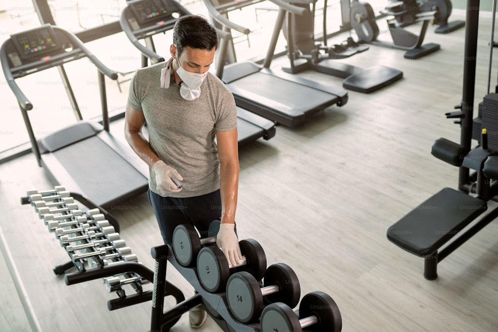 Male athlete wearing face mask and disinfecting hand weights in a gym during COVID-19 epidemic.