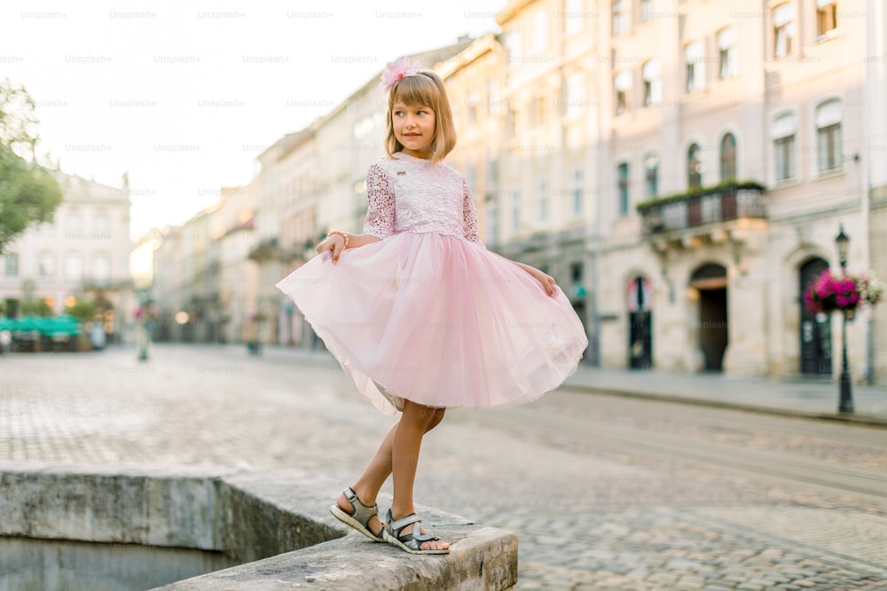 Ritratto a figura intera di moda di una bambina bionda carina, che indossa un vestito rosa e un fiore tra i capelli, in posa per la macchina fotografica, mentre si trova su un'antica fontana di pietra nella vecchia città europea.