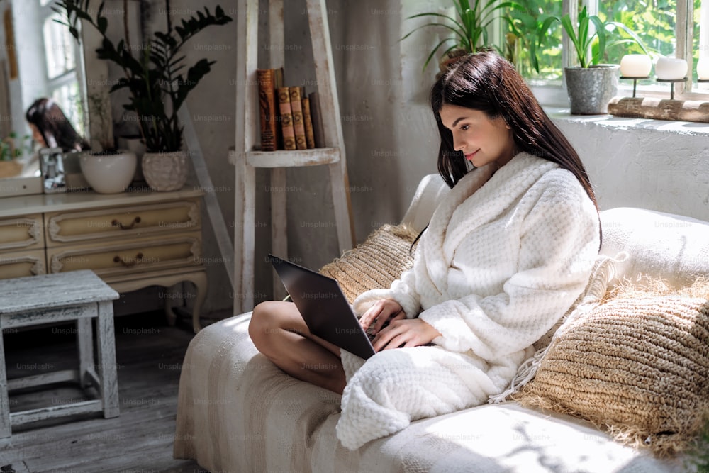 Side view of young adult woman sitting on sofa in bathrobe, using modern laptop computer. girl making smiling face, resting on weekend at home