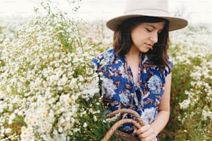 Hermosa muchacha sosteniendo un ramo de margaritas blancas. Verano tranquilo en el campo. Mujer joven con estilo en vestido vintage azul y sombrero que recoge flores silvestres blancas en una canasta de paja en el prado.