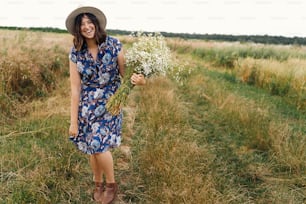 青いビンテージドレスと帽子をかぶったスタイリッシュな若い女性が、夏の牧草地で白い野の花と一緒に歩いています。フィールドでヒナギクの花束で微笑む幸せな美しい女の子。田舎の夏