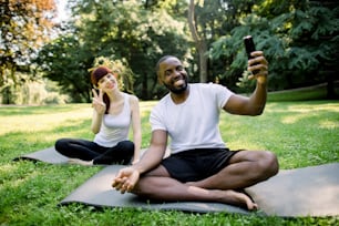Yoga at park. Horizontal shot of funny young multiethnic family couple ready for youga and exercising outdoors, sitting on the fit mats and making photo on smartphone. Concept of healthy lifestyle