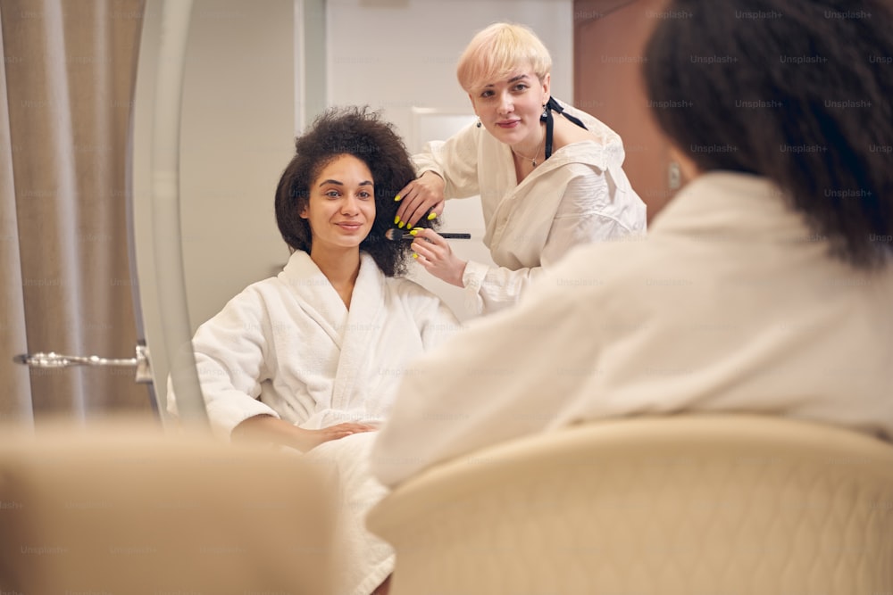 Positive experienced beauty parlor employee touching woman hair while looking at reflection of her work