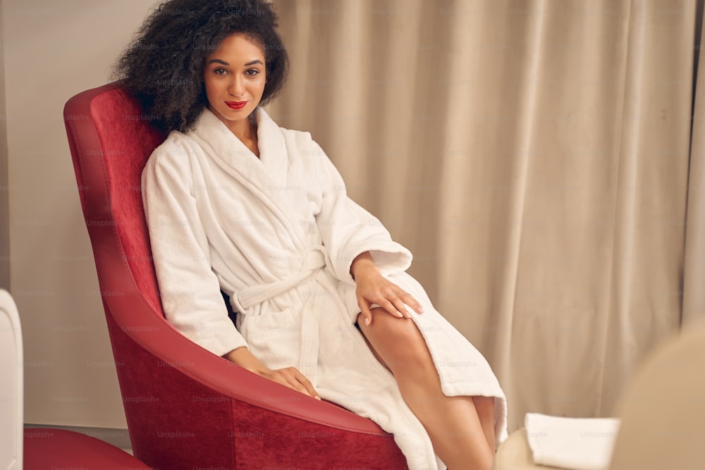 Pleasant curly haired woman resting in convenient red armchair while keeping legs crossed