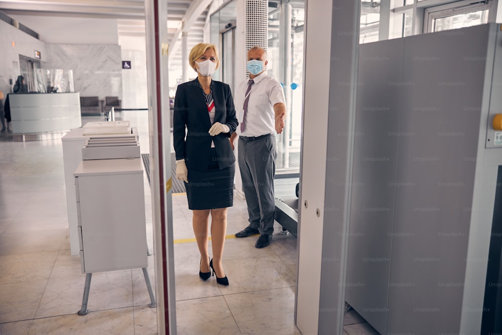 Charming businesswoman in medical mask passing through check-in gate while male airport worker standing behind her