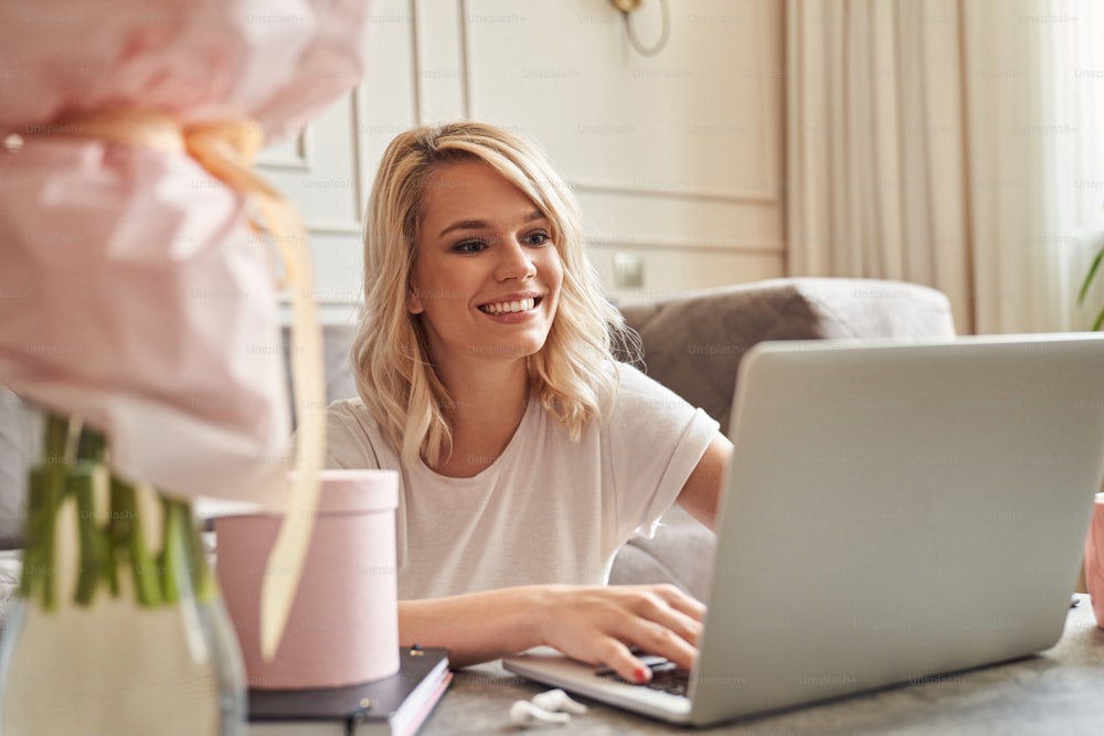 Happy blonde woman sitting at home with lovely present and flowers by her side and smiling while looking at the screen of a laptop