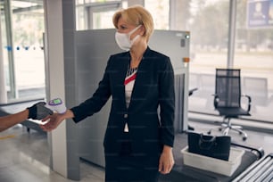 Female airport worker using digital medical thermometer while checking body temperature of elegant businesswoman in medical mask