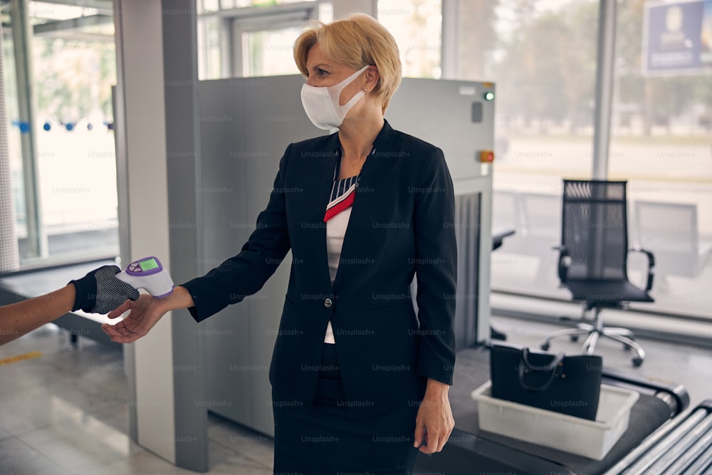 의료 마스크를 쓴 우아한 사업가의 체온을 확인하면서 디지털 체온계를 사용하는 여성 공항 노동자