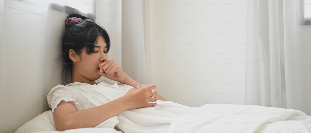 An ill woman drinks water and consuming a pill while lying on the bed in the bedroom.
