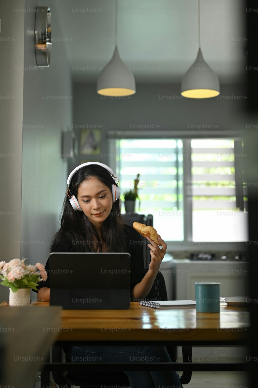 A beautiful woman is eating a croissant while using a computer tablet at the wooden working desk.