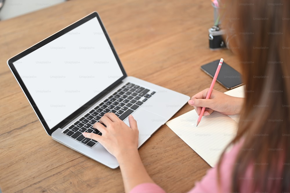 Uma imagem recortada de uma mulher está usando um laptop de computador de tela branca em branco na mesa de madeira.