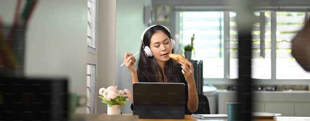 A beautiful woman is eating a croissant while using a computer tablet at the wooden working desk.