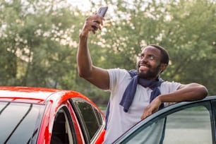 Young smiling handsome African American man in white t-shirt, standing near his new luxury red car and taking selfie pictures with his smartphone. Happy satisfied new owner of the car outdoors.