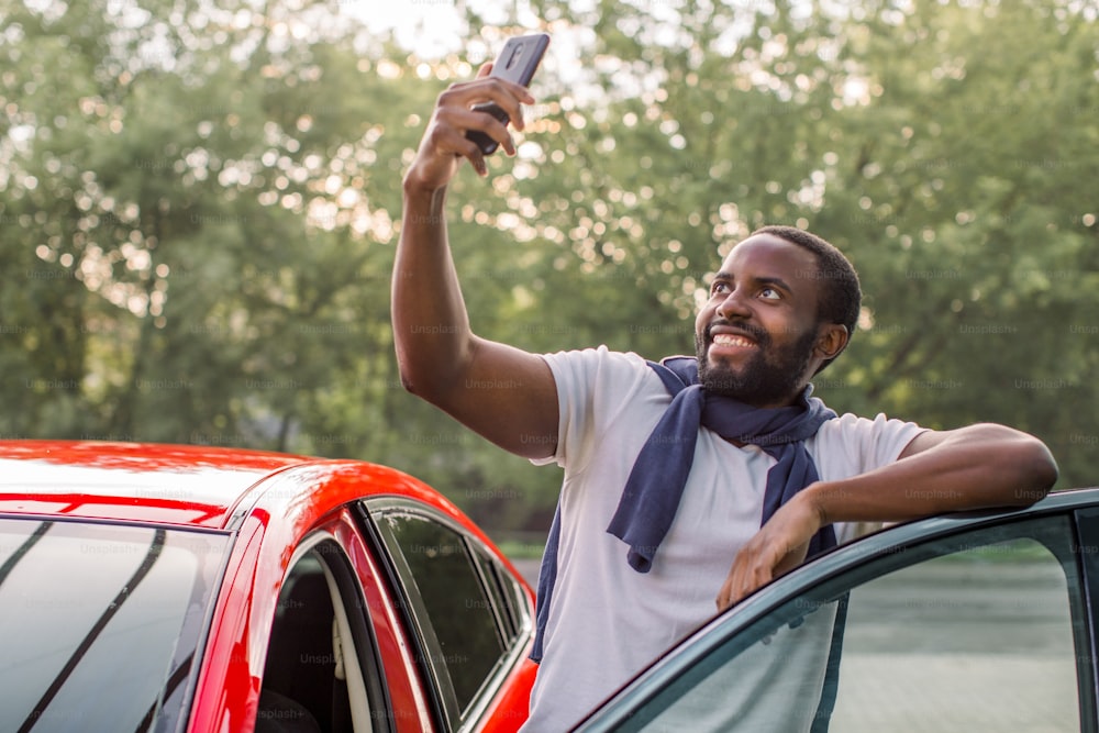 Young smiling handsome African American man in white t-shirt, standing near his new luxury red car and taking selfie pictures with his smartphone. Happy satisfied new owner of the car outdoors.